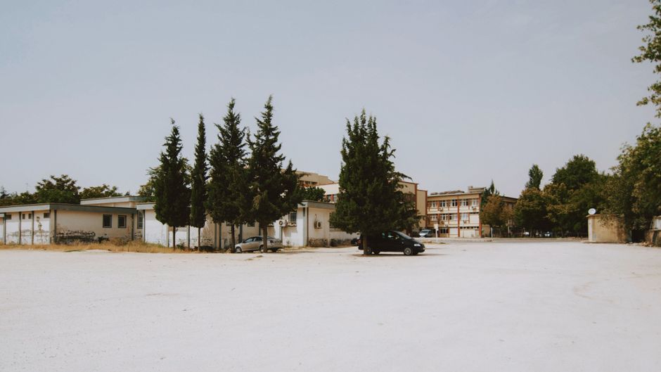 Wide shot of an empty urban lot with buildings and tall trees under a clear sky.